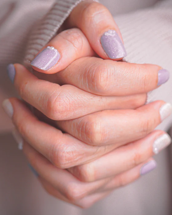 Mains aux ongles lilas jointes avec douceur sur un drapé rose pâle